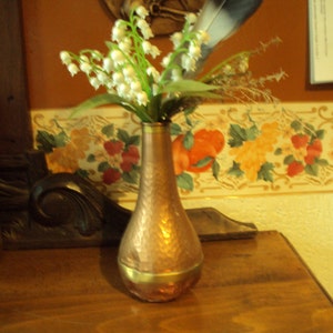 Può includere: Un vaso di rame con finitura martellata che contiene un mazzo di mughetti bianchi. Il vaso ha un bordo dorato e si trova su una superficie di legno.