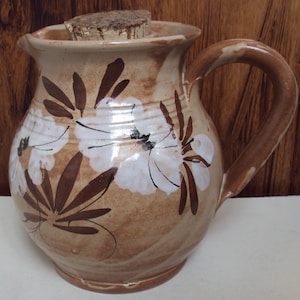 May include: A brown ceramic jug with a cork lid and white floral design. The jug has a brown handle and a slightly rounded body.