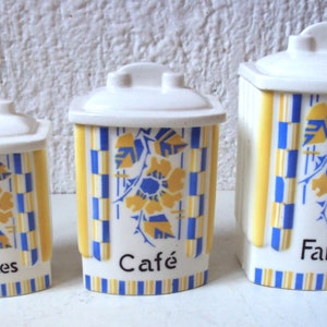 Op de afbeelding: Drie witte keramische potten met blauwe en gele geometrische patronen en bloemmotieven. De potten zijn gelabeld met "Epices", "Cafe" en "Farine".