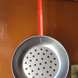 Peut inclure: Une passoire en métal argenté avec une poignée rouge suspendue à un crochet. La passoire est de forme circulaire et présente un motif de petits trous.