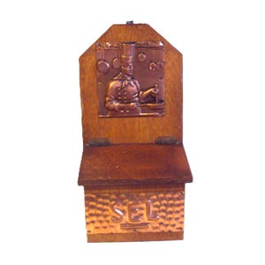 Puede incluir: Un salero de madera con frente de cobre y relieve de cobre de un chef. La caja tiene una tapa con bisagras y un relieve decorativo de cobre de un chef cocinando en una cocina. La palabra "SAL" está grabada en la parte delantera.