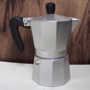 Peut inclure: Une cafetière à espresso en aluminium argenté avec une poignée noire. La cafetière a une base ronde octogonale et un bec verseur sur le côté.