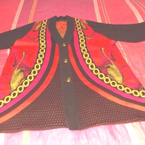 Peut inclure: Un cardigan noir avec un motif de chaîne rouge, rose et or. Le cardigan a un col en V et une fermeture à boutons.