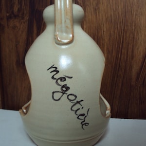 Op de afbeelding: Een beige keramische fles met een handvat en een uitsparing aan de zijkant. De fles heeft het woord "onderhandelen" in het zwart erop geschreven.