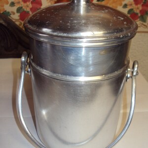 May include: A vintage, brushed aluminum ice bucket with a lid and a handle. The bucket has a cylindrical shape with a slightly flared base. The lid has a small knob on top. The handle is a curved metal bar attached to the sides.