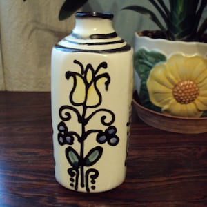 Peut inclure: Un vase en céramique blanche avec une fleur jaune et un motif floral noir et bleu. Le vase a une base arrondie et un col étroit.