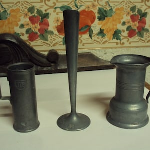 May include: Three pewter items: a tall, slender vase, a small pitcher with a handle, and a cylindrical mug with a handle. The mug has the letters "RP" embossed on the front.