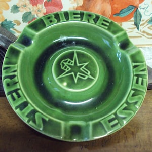 Puede incluir: Cenicero de cerámica verde con las palabras "BIERE" y "ESSEN" escritas alrededor del borde. El centro del cenicero presenta una estrella con una imagen estilizada de una jarra de cerveza.