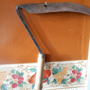 May include: A rusty, curved metal blade with a wooden handle. The blade is attached to the handle with a metal rivet. The blade is hanging on a wall with a floral wallpaper border.