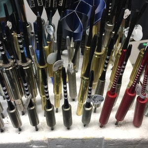 May include: A collection of darts with different colored shafts and flights. The shafts are made of metal and the flights are made of plastic. The darts are arranged in a row on a white surface.