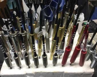 May include: A collection of darts with different colored shafts and flights. The shafts are made of metal and the flights are made of plastic. The darts are arranged in a row on a white surface.
