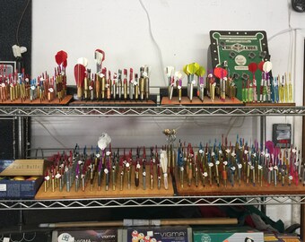 May include: Two shelves filled with a variety of colorful dart shafts. The shafts are arranged in rows on wooden boards. The top shelf has a dart game board with the text "The One and Only Cage".