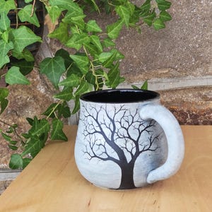 May include: A ceramic mug with a rounded body and a thick handle. The exterior features a light gray finish with a hand-painted black silhouette of a leafless tree with spreading branches. The interior of the mug is finished in a solid black glaze.