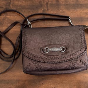May include: A dark brown textured crossbody bag with a flap closure. The bag features decorative whipstitch detailing along the edges and a central silver-toned metal hardware piece with an engraved floral pattern and two circular rings.