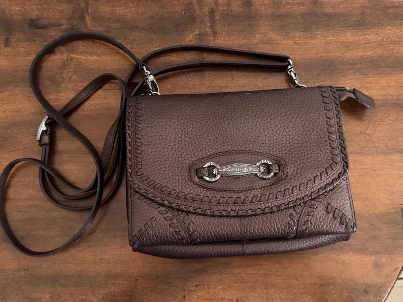 May include: A dark brown textured crossbody bag with a flap closure. The bag features decorative whipstitch detailing along the edges and a central silver-toned metal hardware piece with an engraved floral pattern and two circular rings.
