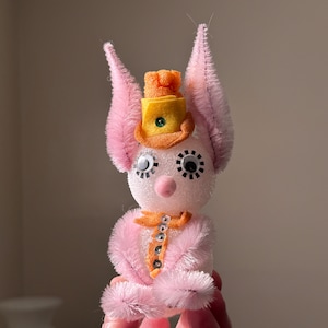 May include: A white, fuzzy, handmade toy with pink fur, large black eyes, and a yellow and orange hat. The toy is sitting on a hand.