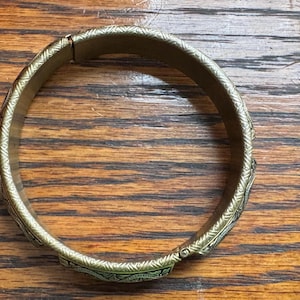 May include: A circular brass-toned metal bangle bracelet featuring a continuous engraved geometric pattern along its outer surface. The bracelet has a hinged opening mechanism with a visible clasp, showcasing a metallic finish and intricate detailing.