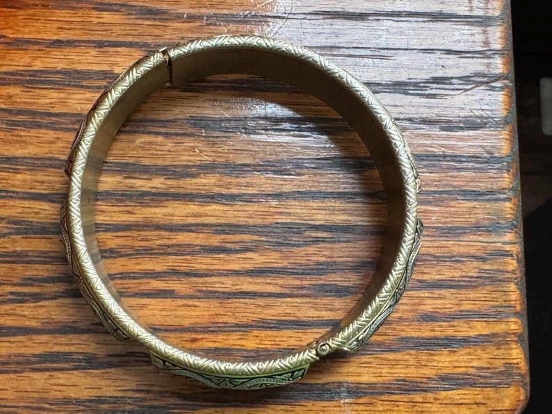 May include: A circular brass-toned metal bangle bracelet featuring a continuous engraved geometric pattern along its outer surface. The bracelet has a hinged opening mechanism with a visible clasp, showcasing a metallic finish and intricate detailing.