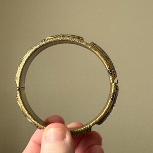 May include: A circular gold-toned metal bangle bracelet featuring a repeating engraved pattern along the outer surface. The bracelet has a hinged opening mechanism on one side, allowing it to be opened and closed securely around the wrist.