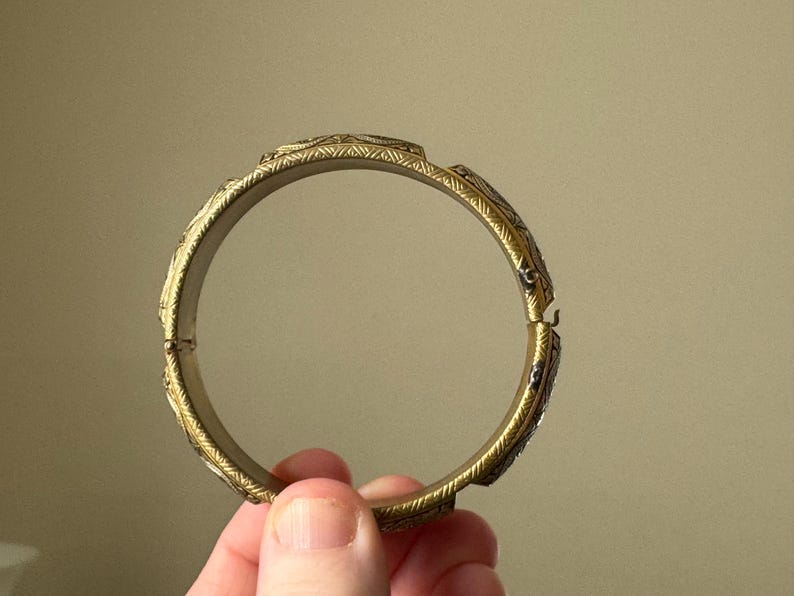 May include: A circular gold-toned metal bangle bracelet featuring a repeating engraved pattern along the outer surface. The bracelet has a hinged opening mechanism on one side, allowing it to be opened and closed securely around the wrist.