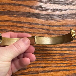 May include: A gold-toned metal hinged bangle bracelet with a smooth, curved interior surface and a visible mechanical hinge mechanism at the center. The bracelet features decorative end caps and a polished finish, showing a semi-circular shape.