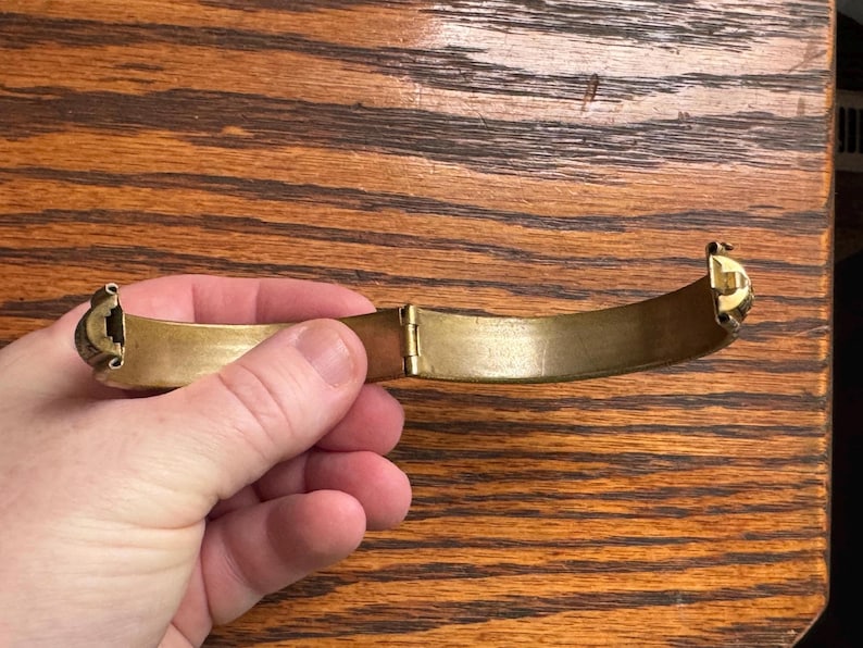 May include: A gold-toned metal hinged bangle bracelet with a smooth, curved interior surface and a visible mechanical hinge mechanism at the center. The bracelet features decorative end caps and a polished finish, showing a semi-circular shape.