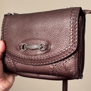 May include: A dark brown, textured leather crossbody bag featuring a metallic finish. The front flap is adorned with a decorative, engraved silver-toned metal hardware piece and intricate braided leather stitching along the curved edges.
