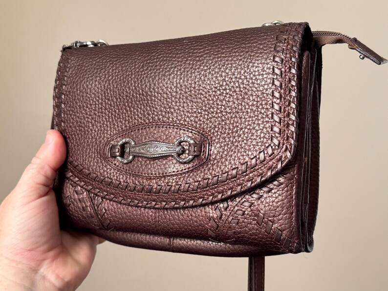 May include: A dark brown, textured leather crossbody bag featuring a metallic finish. The front flap is adorned with a decorative, engraved silver-toned metal hardware piece and intricate braided leather stitching along the curved edges.