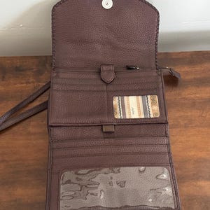 May include: A dark brown pebbled leather wallet with a fold-over flap and a snap closure. The interior features multiple card slots, a central zippered compartment, a clear ID window, and a large transparent pocket at the bottom for documents.