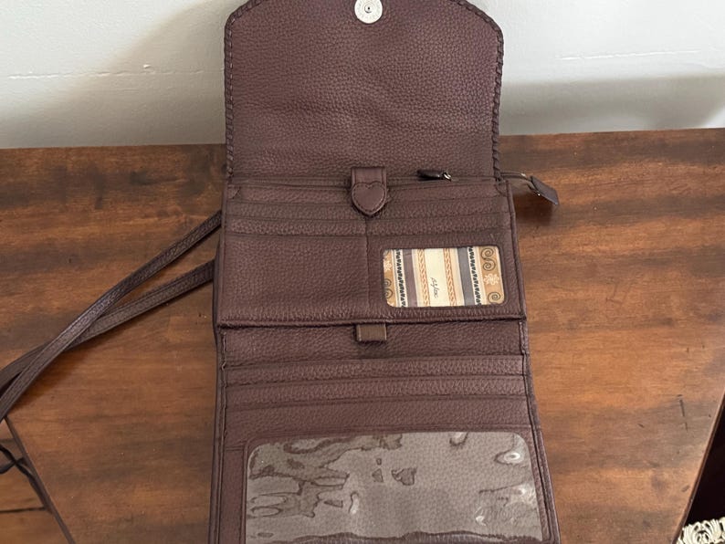 May include: A dark brown pebbled leather wallet with a fold-over flap and a snap closure. The interior features multiple card slots, a central zippered compartment, a clear ID window, and a large transparent pocket at the bottom for documents.