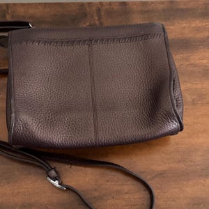 May include: A dark brown leather crossbody bag with a pebbled texture, a vertical center seam, and a decorative whipstitch detail along the top flap. The bag features a thin, adjustable shoulder strap with a silver-toned metal buckle hardware.