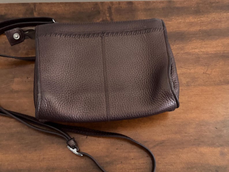 May include: A dark brown leather crossbody bag with a pebbled texture, a vertical center seam, and a decorative whipstitch detail along the top flap. The bag features a thin, adjustable shoulder strap with a silver-toned metal buckle hardware.