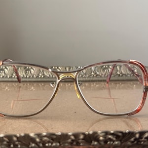 May include: Vintage eyeglasses with a metal frame and clear lenses. The frames have a gold bridge and reddish-brown accents on the upper rims. The glasses are set on a decorative silver tray.