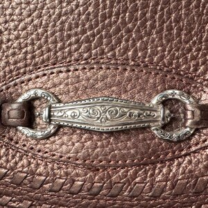 May include: A metallic bronze-colored textured leather surface featuring a central silver-toned metal hardware piece. The hardware consists of an elongated, ornate, engraved bar connected to two circular rings, all secured by leather loops on the material.