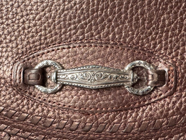 May include: A metallic bronze-colored textured leather surface featuring a central silver-toned metal hardware piece. The hardware consists of an elongated, ornate, engraved bar connected to two circular rings, all secured by leather loops on the material.