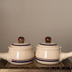 May include: Two cream-colored ceramic soup crocks with single handles and matching lids. Each lid features a dark brown wooden knob. The base of each crock and the lid are decorated with a horizontal stripe pattern in dark blue and deep red.