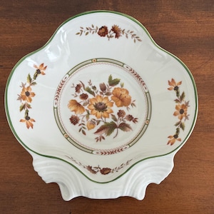 May include: White ceramic serving dish with a scalloped edge and a green trim. The dish features a central floral design in shades of orange, brown, and yellow, surrounded by a decorative border. Additional floral accents are placed around the rim.