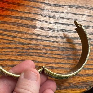 May include: A gold-toned hinged bangle bracelet featuring a repeating etched pattern along the outer surface. The bracelet has a curved, semi-circular shape with a visible metal hinge mechanism and a clasp closure at the ends of the band.