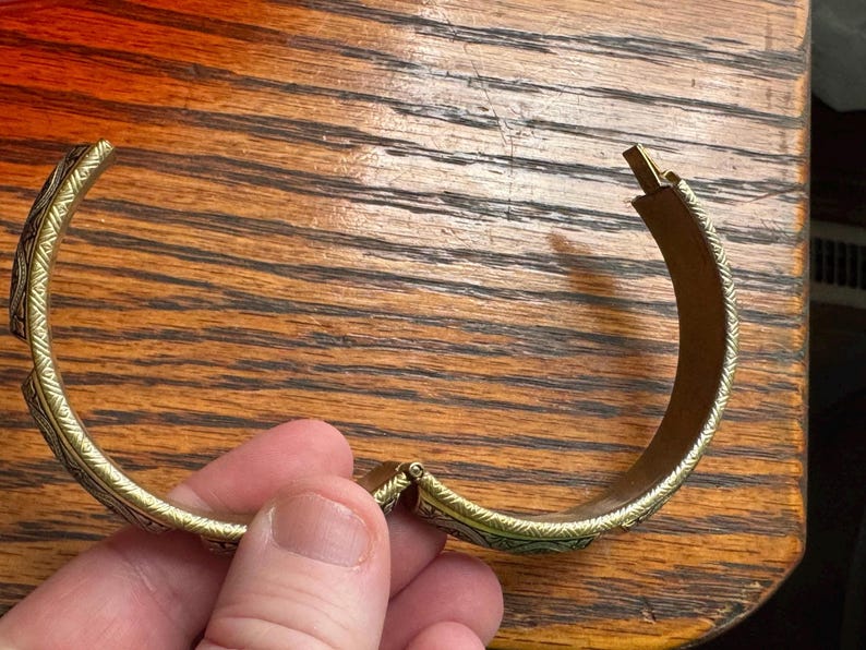 May include: A gold-toned hinged bangle bracelet featuring a repeating etched pattern along the outer surface. The bracelet has a curved, semi-circular shape with a visible metal hinge mechanism and a clasp closure at the ends of the band.