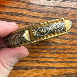 May include: A hinged metal bracelet featuring a two-tone design with gold-colored edges and a central silver-colored panel. The silver section is decorated with repeating embossed diamond-shaped patterns containing floral motifs and dark accents.
