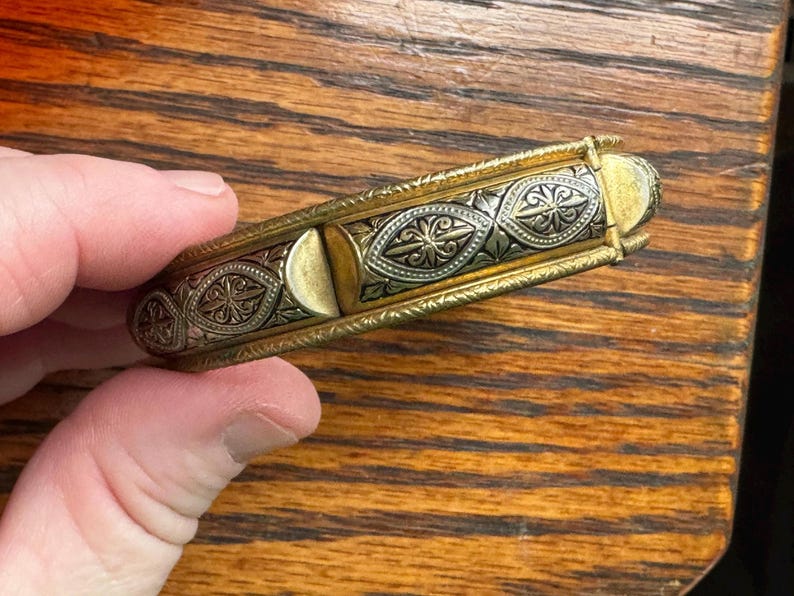 May include: A hinged metal bracelet featuring a two-tone design with gold-colored edges and a central silver-colored panel. The silver section is decorated with repeating embossed diamond-shaped patterns containing floral motifs and dark accents.
