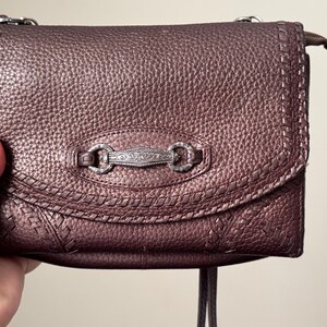 May include: A metallic bronze-toned shoulder bag with a pebbled texture, featuring a decorative silver-colored metal bar with intricate floral engravings at the center of the flap, accented by stitched detailing along the edges and a zipper closure.