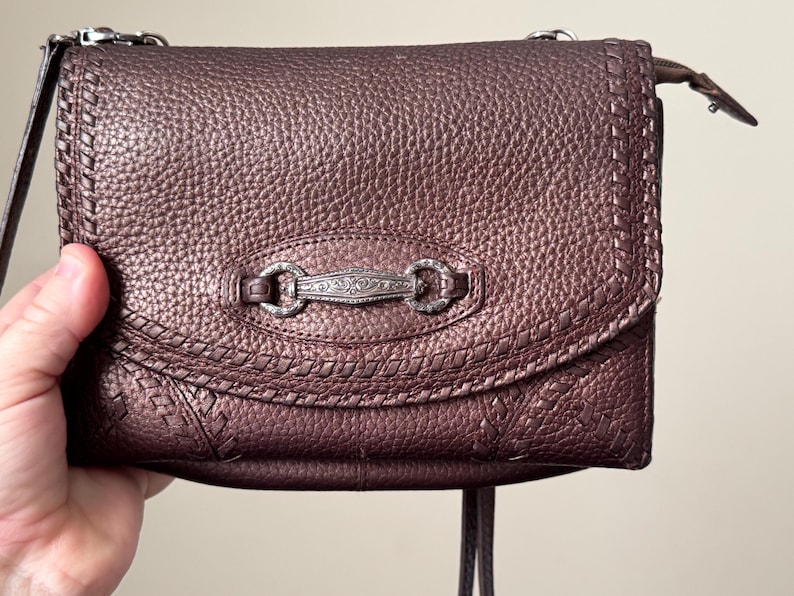 May include: A metallic bronze-toned shoulder bag with a pebbled texture, featuring a decorative silver-colored metal bar with intricate floral engravings at the center of the flap, accented by stitched detailing along the edges and a zipper closure.