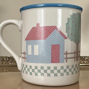 May include: White ceramic mug with a blue rim and handle. The mug features a cross-stitch design of a house, tree, and fence in red, blue, and green. The mug is a standard size for coffee or tea.