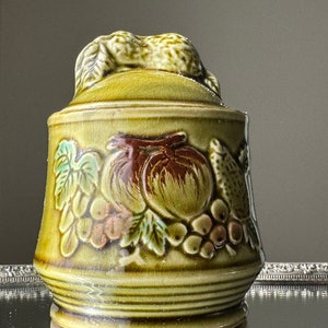 May include: A green ceramic jar with a lid featuring a raised fruit design. The jar has a brown, green, and pink fruit design with leaves.