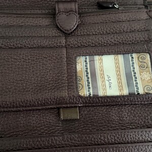 May include: A dark brown textured leather wallet featuring multiple card slots, a heart-shaped snap closure, and a clear ID window displaying a patterned fabric lining with the text Brighton printed in a script font on the interior panel.