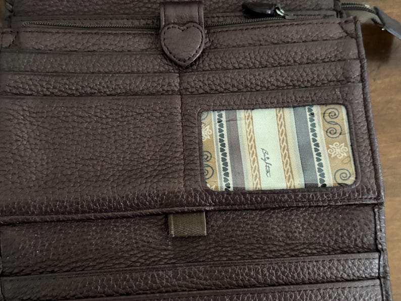 May include: A dark brown textured leather wallet featuring multiple card slots, a heart-shaped snap closure, and a clear ID window displaying a patterned fabric lining with the text Brighton printed in a script font on the interior panel.
