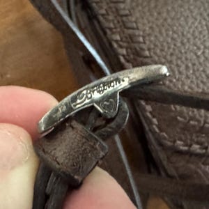 May include: A silver-toned metal toggle clasp featuring the engraved word Brighton and a small heart symbol. The clasp is attached to a dark brown textured leather strap, which is part of a larger brown leather item with a visible stitched pattern.