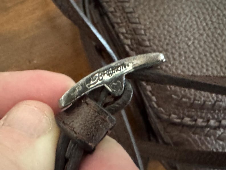 May include: A silver-toned metal toggle clasp featuring the engraved word Brighton and a small heart symbol. The clasp is attached to a dark brown textured leather strap, which is part of a larger brown leather item with a visible stitched pattern.
