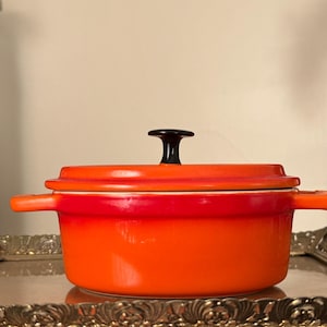 May include: A small, orange enameled cast iron pot with a lid and two side handles. The lid has a black handle. The pot is sitting on a decorative silver tray. The pot is likely used for cooking.